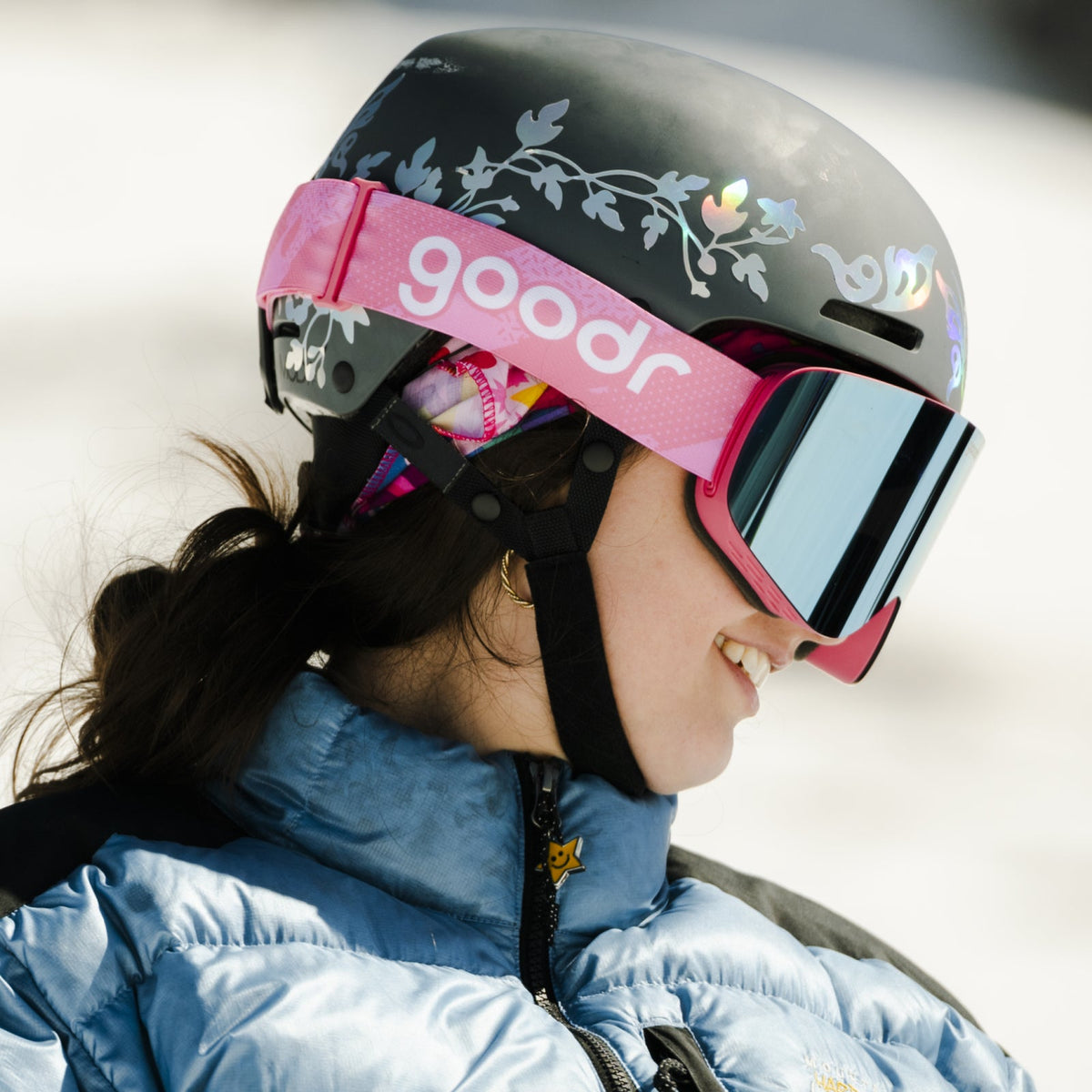 Smiling person in 90s Ski Movie Villain goodr pink mirrored goggles and a black floral ski helmet.