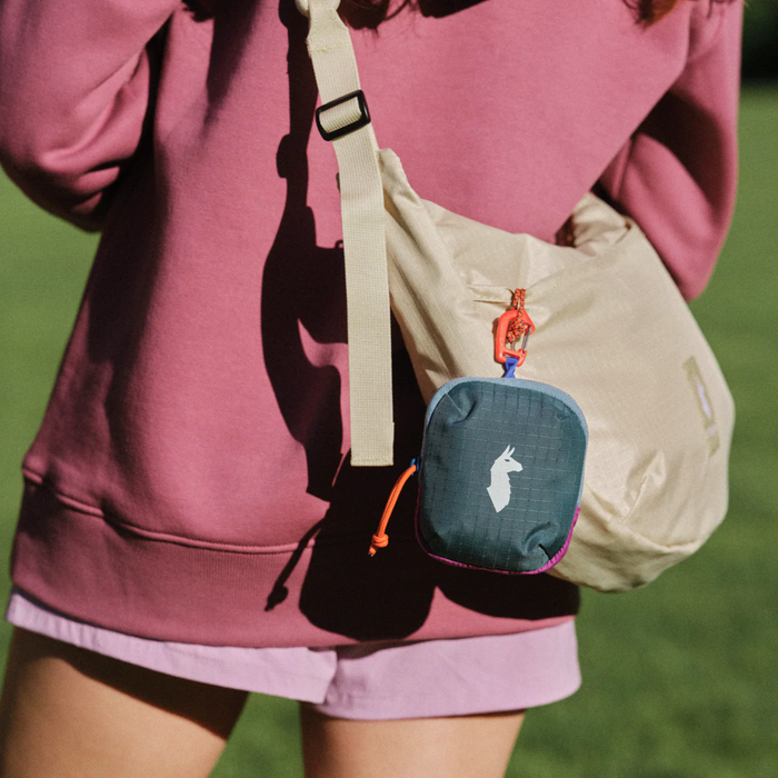 Close-up of a person carrying a beige shoulder bag with a dark teal Cotopaxi Allpita Mini Bag attached, featuring colorful details.