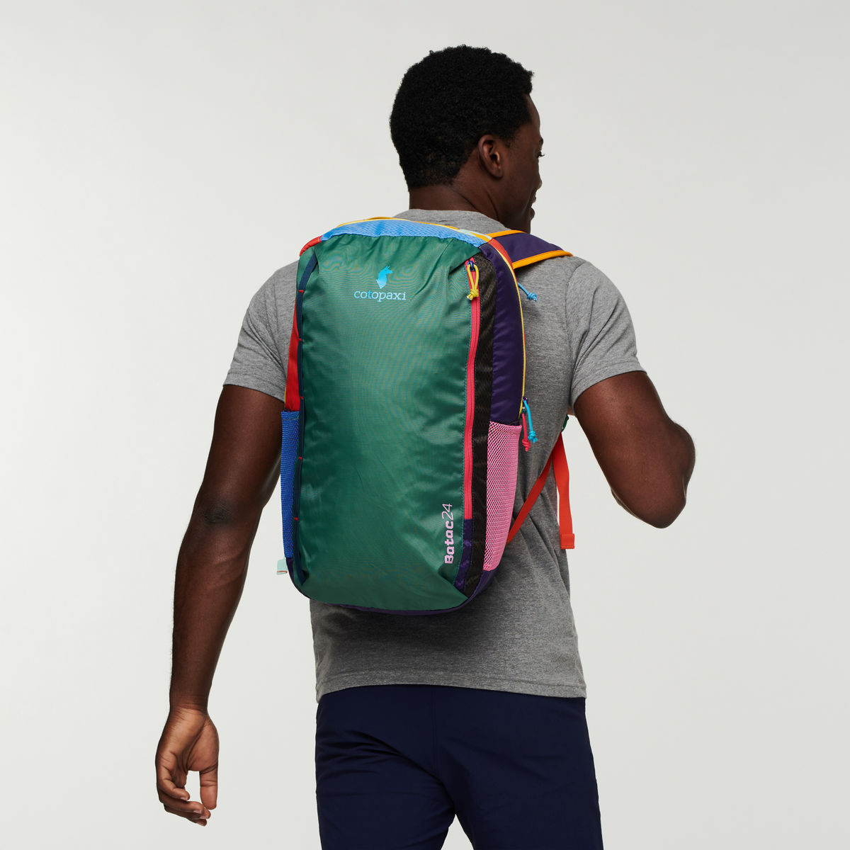 Man wearing a colorful Cotopaxi Batac 24L backpack, green with pink, blue, red, and purple accents, seen from behind.