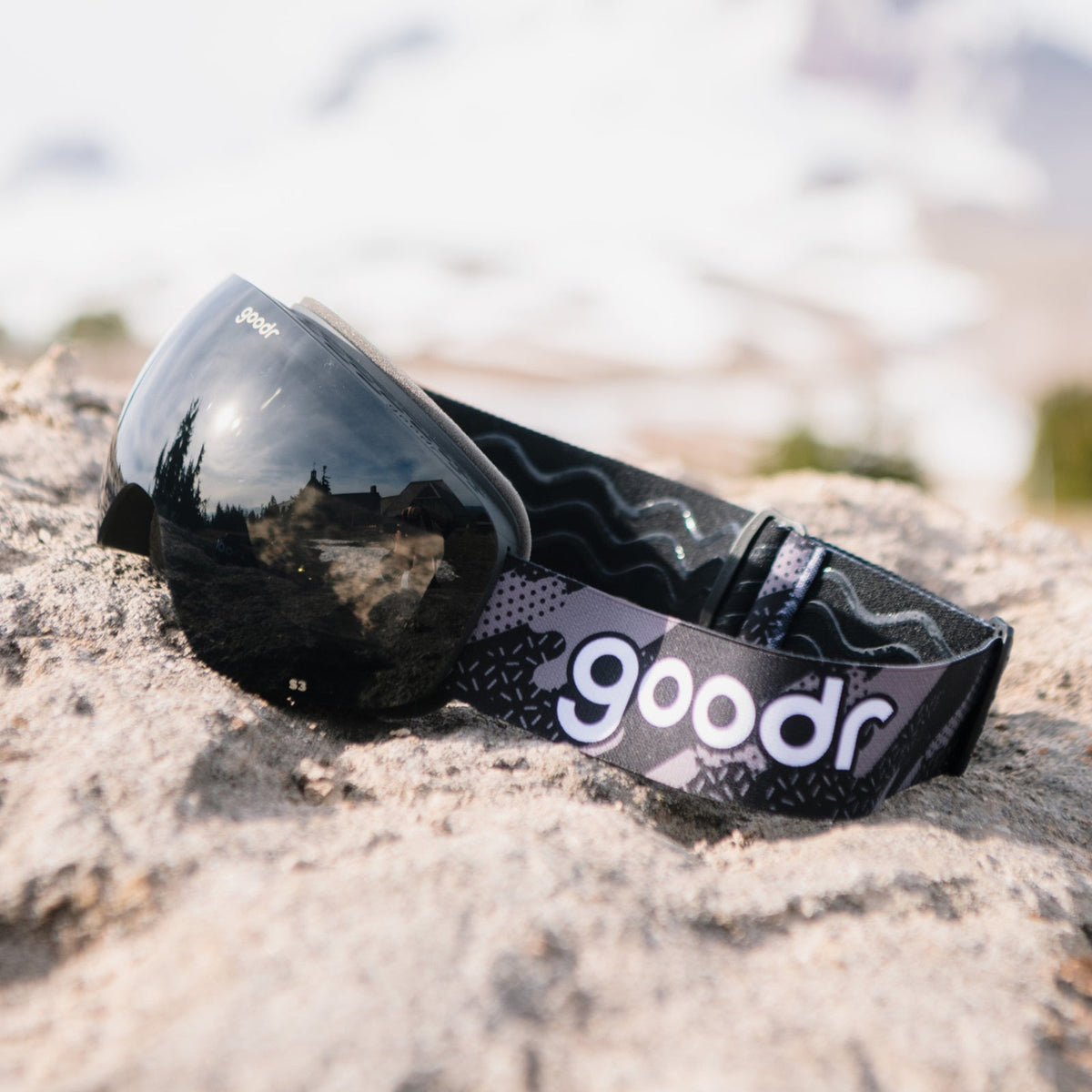 Beware of Shadow Ridge | goodr ski goggles with black lenses and patterned strap on a rocky surface, reflecting trees and sky.