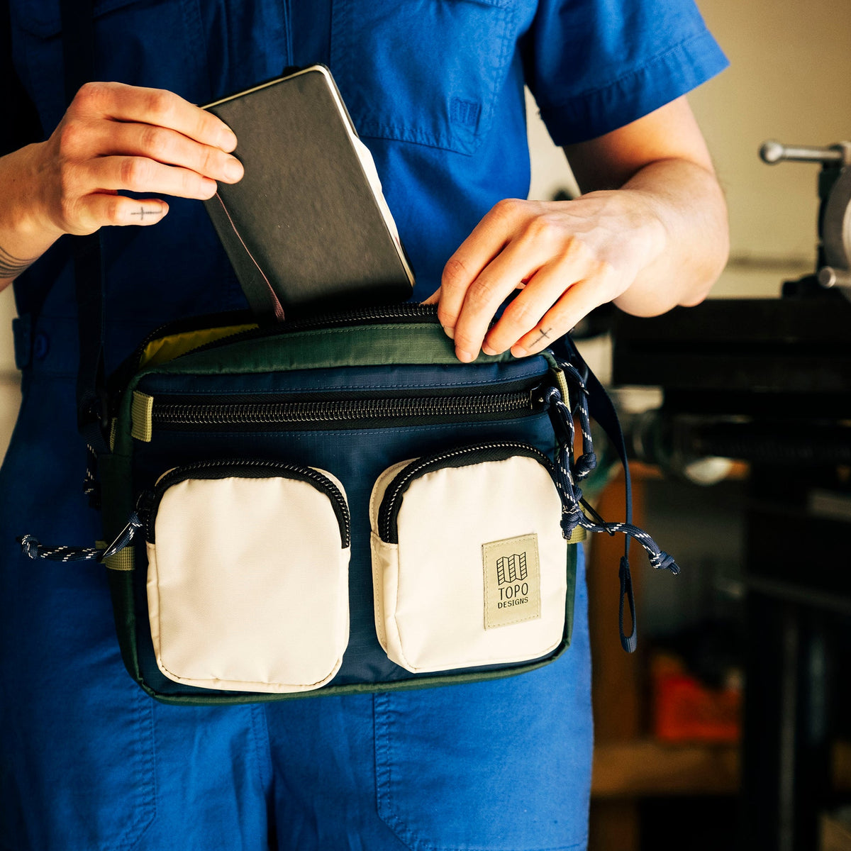 Person in blue putting a notebook into a navy and green Topo Designs Block Bag with two cream zippered front pockets.