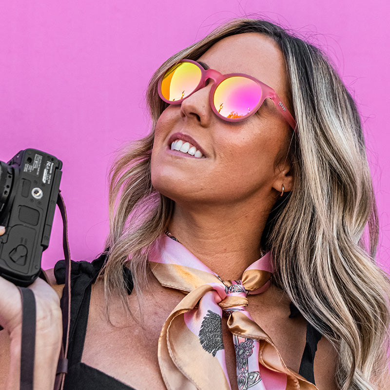 Woman wearing pink goodr "Influencers Pay Double" sunglasses, holding a camera, against a vibrant pink background.