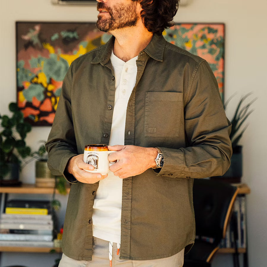 Man in an olive green Topo Designs Men's Desert Shirt, worn over a white henley, holding a coffee mug.