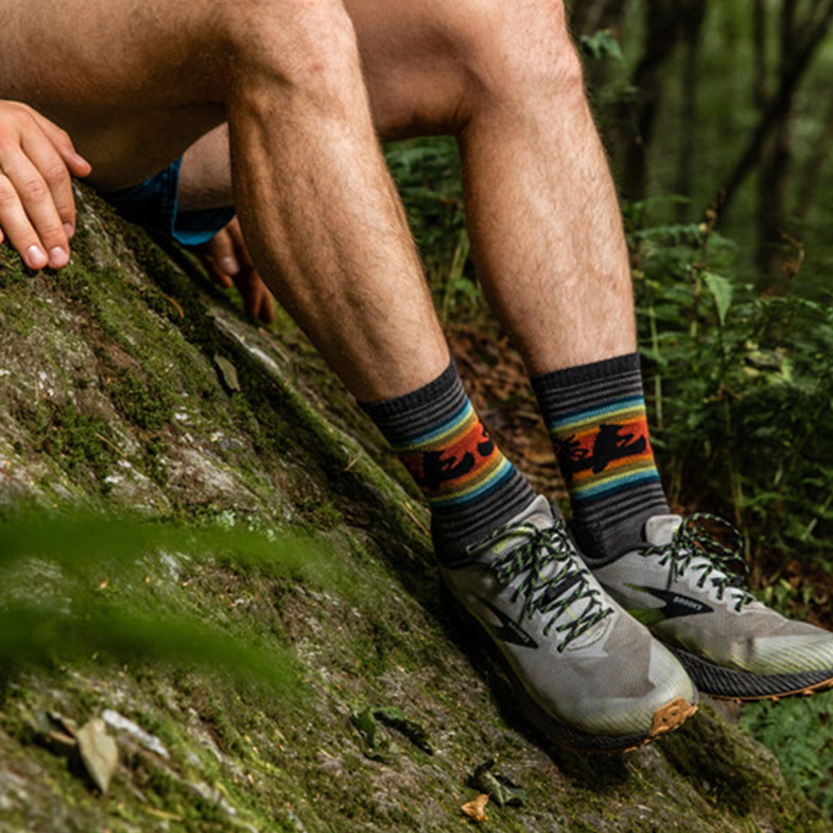 Men's Willoughby Micro Crew | Darn Tough socks, black with colorful stripes and bear silhouettes, worn with trail shoes on a mossy rock.