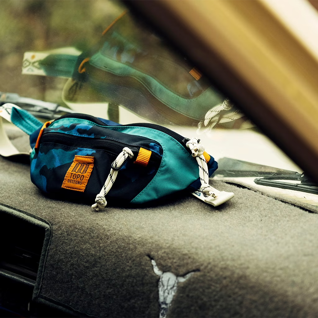 Mountain Waist Pack by Topo Designs, in a teal, blue, and black camo pattern, resting on a gray car dashboard.