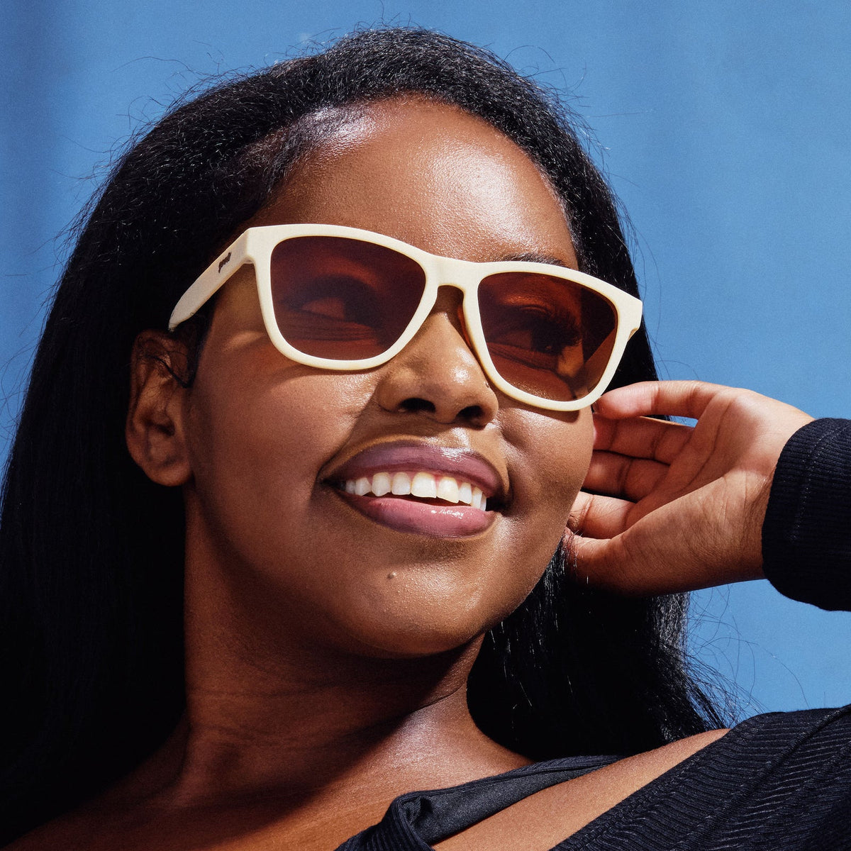 Dark-skinned woman smiling in 'Toasted Marshmallow Magic | goodr' off-white square sunglasses, blue sky background.