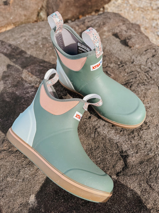 XTRATUF Women's Ankle Deck Boots in Garden Green, with peach and light blue accents, resting on textured rocks.