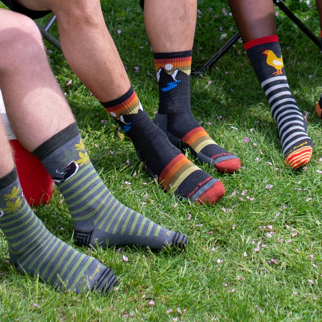 Men's Sunset Ridge Micro Crew Darn Tough socks with mountain and sunset design, worn on green grass with other patterned socks.