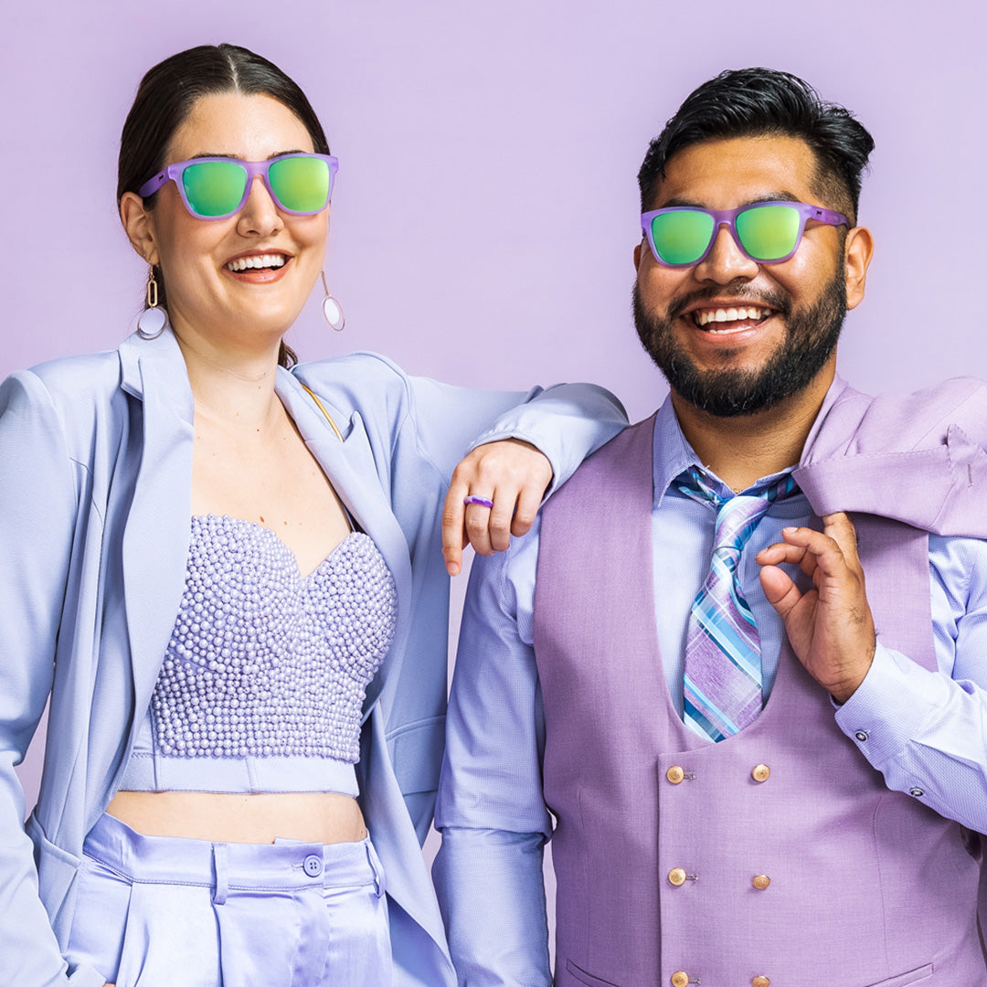 Man and woman wearing lilac outfits and purple framed goodr Lilac It Like That!!! sunglasses with green reflective lenses.