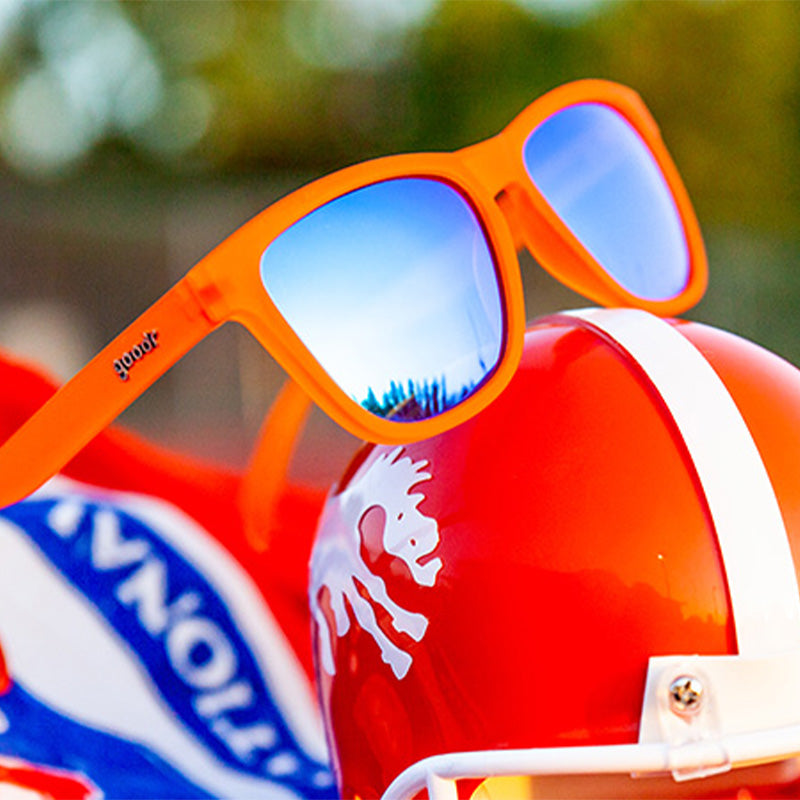 Orange goodr "Donkey Goggles" sunglasses with blue reflective lenses resting on a red football helmet with a white logo.