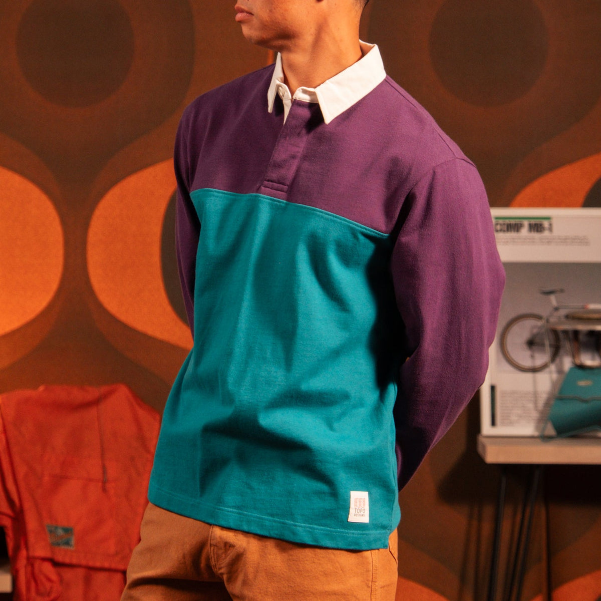 Men's Topo Designs Mountain Rugby Shirt in a vibrant purple and teal color block, worn by a man with brown pants.