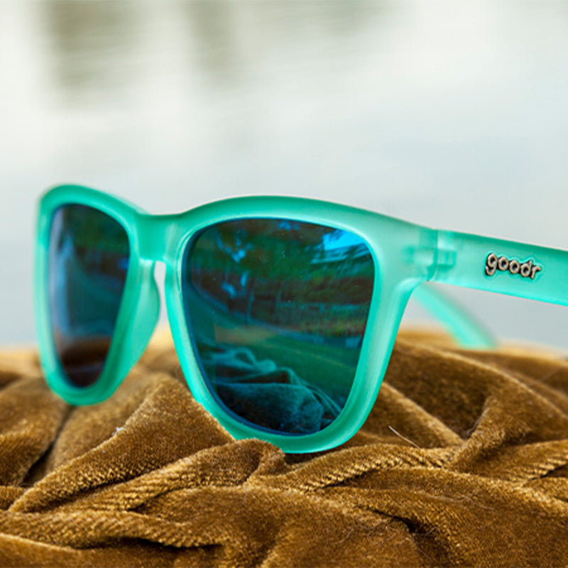 Close-up of Nessy's Midnight Orgy goodr sunglasses, featuring a turquoise frame and blue mirrored lenses on a textured brown surface.