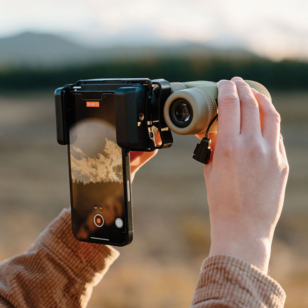 Hands using Nocs Provisions Photo Rig Smartphone Adapter on a monocular, capturing a distant natural landscape on phone screen.