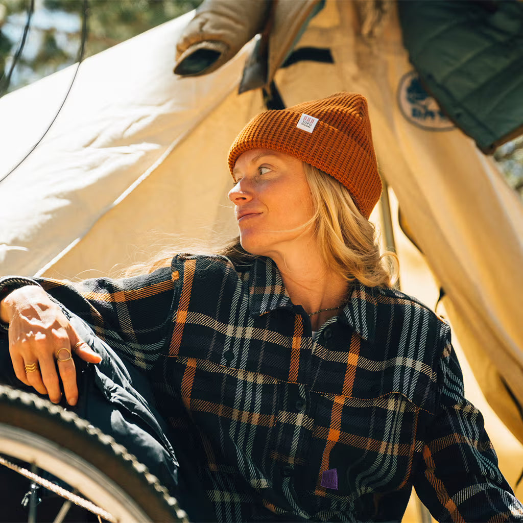 Woman wearing an orange Topo Designs Waffle Knit Beanie and a plaid shirt, sitting inside a tent with a bicycle.