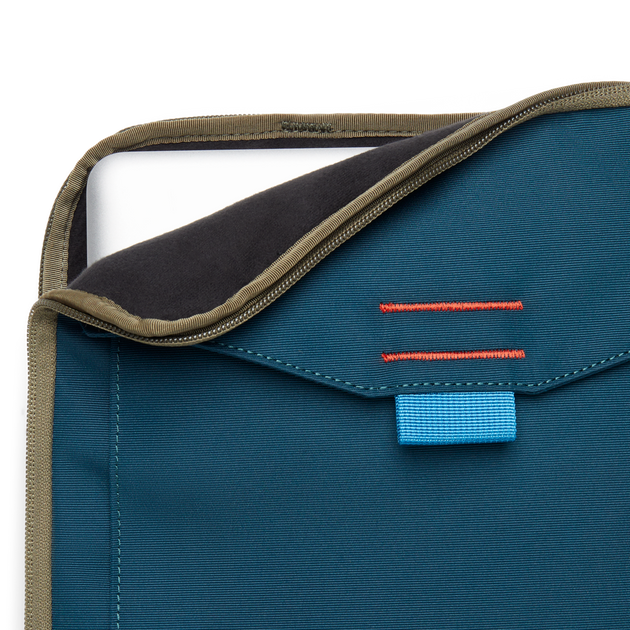 Close-up of a blue Cotopaxi Cada Dia 16" Laptop Sleeve with a laptop partially inserted, showing khaki zipper and blue tag.