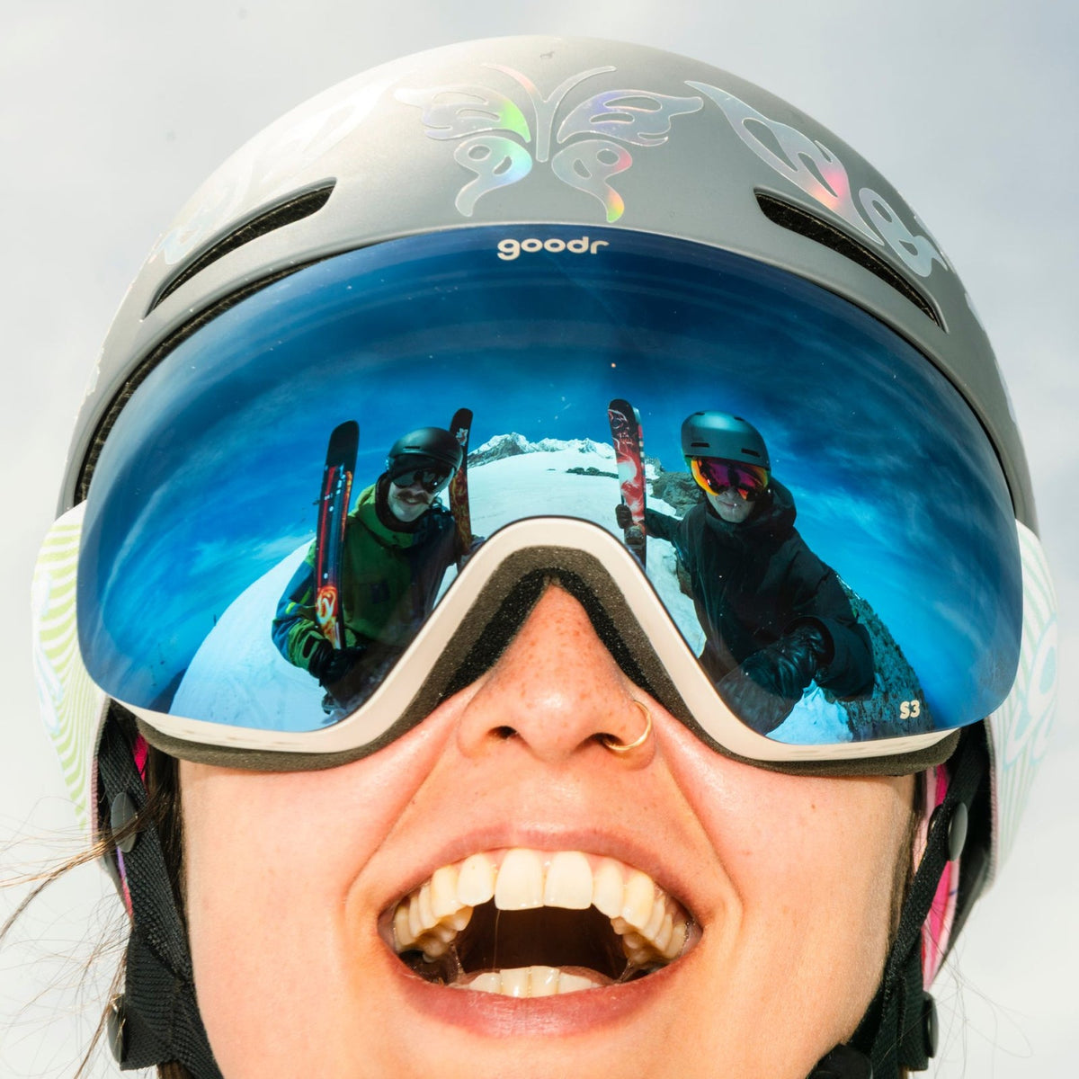 A skier wearing goodr Abominable Snow Icon goggles that reflect two skiers holding skis on a snowy mountain.