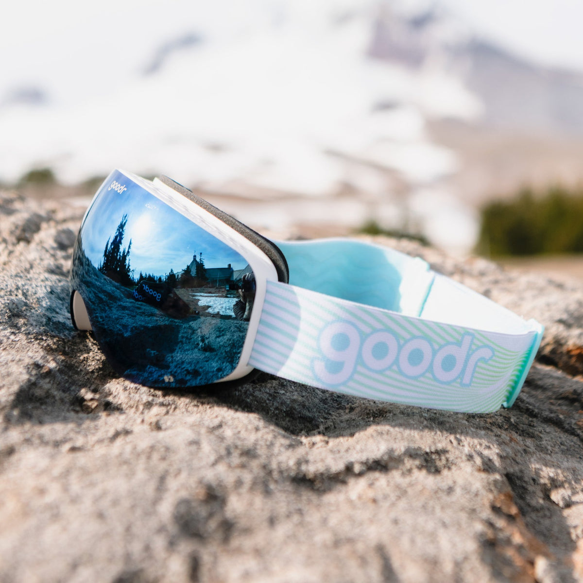Abominable Snow Icon goodr ski goggles with blue mirrored lenses reflecting a snowy lodge and trees, resting on a rock.