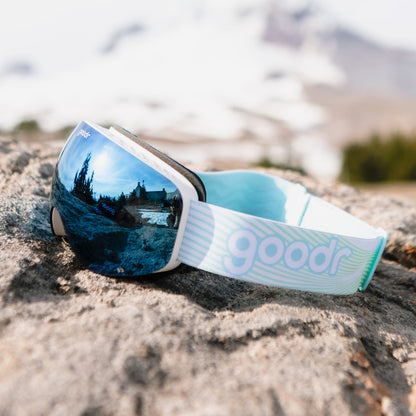 Abominable Snow Icon goodr ski goggles with blue mirrored lenses reflecting a snowy lodge and trees, resting on a rock.