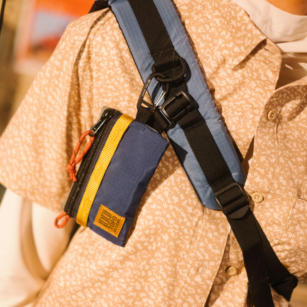 Topo Designs Mountain Accessory Bag in blue and yellow attached to a backpack strap on a person wearing a patterned shirt.