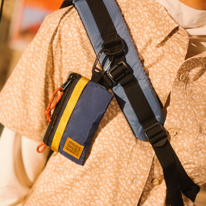 Topo Designs Mountain Accessory Bag in blue and yellow attached to a backpack strap on a person wearing a patterned shirt.