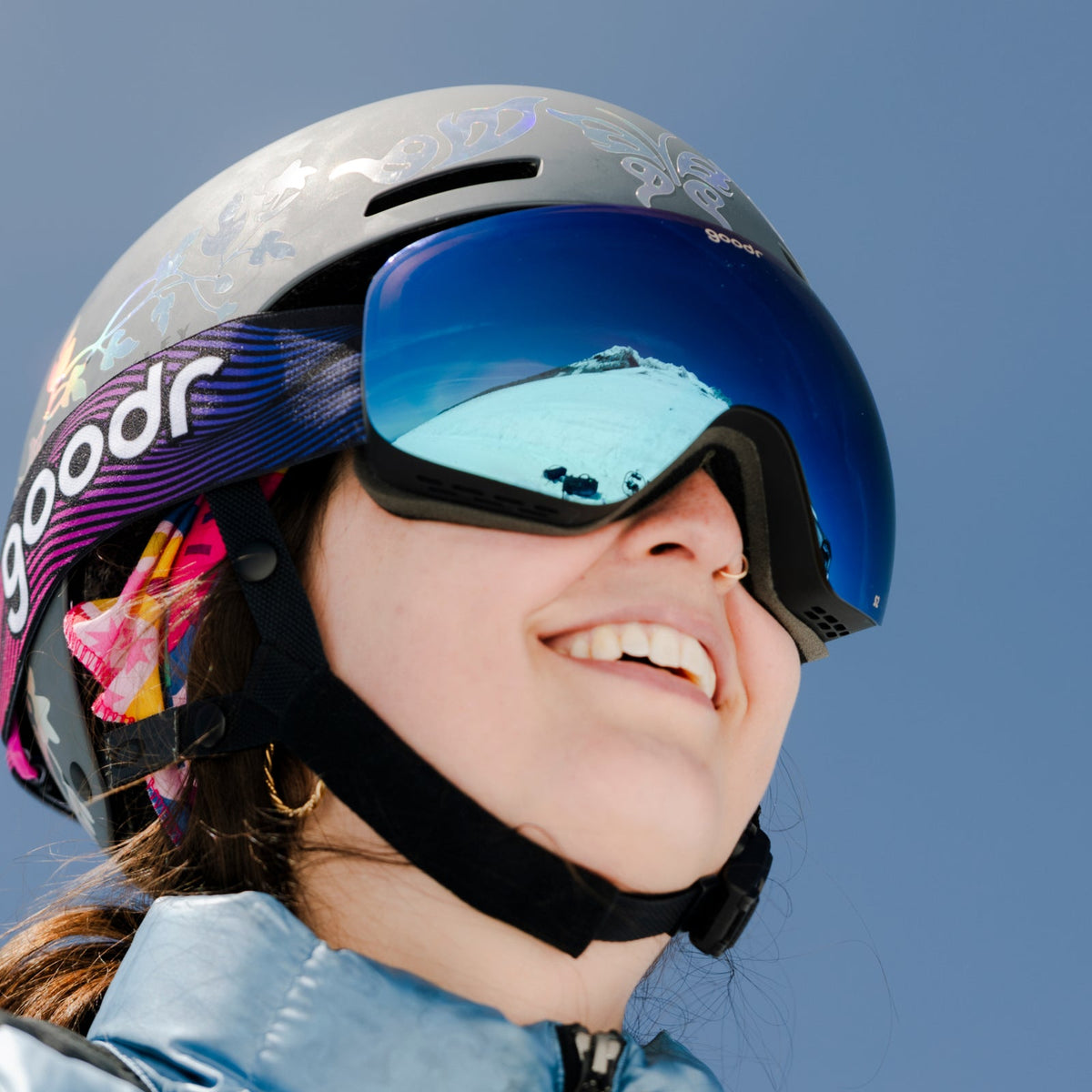 A smiling person wearing an iridescent grey ski helmet and goodr blue mirrored goggles, reflecting a snowy mountain. Alpine State of Mind | goodr.