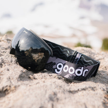 Beware of Shadow Ridge | goodr ski goggles with black lenses and patterned strap on a rocky surface, reflecting trees and sky.