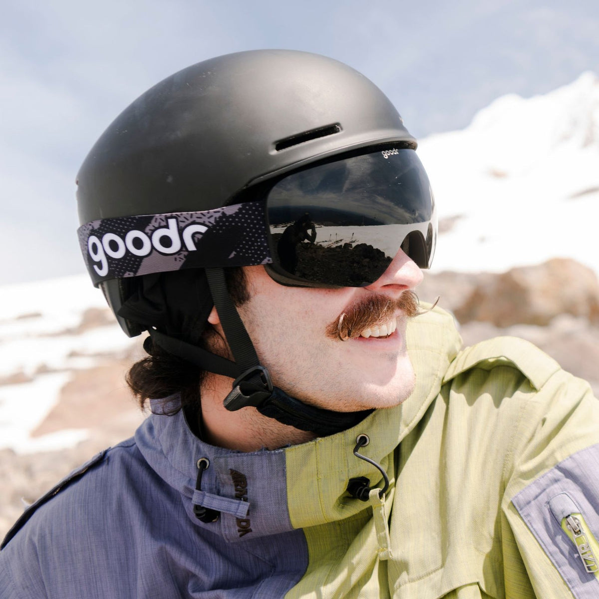 Man in black goodr ski helmet and dark goggles with mustache, looking right at snowy mountain vista. Product: Beware of Shadow Ridge | goodr.