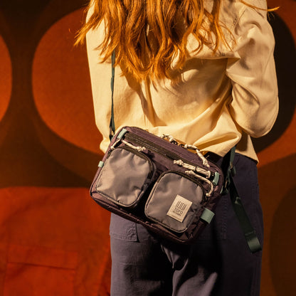 Topo Designs Block Bag in dark purple/grey worn across a person's body from behind, showing front pockets and cord details.
