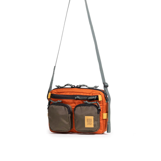 Topo Designs Block Bag in vibrant orange with olive green pockets and a grey strap, shown hanging against a white background.