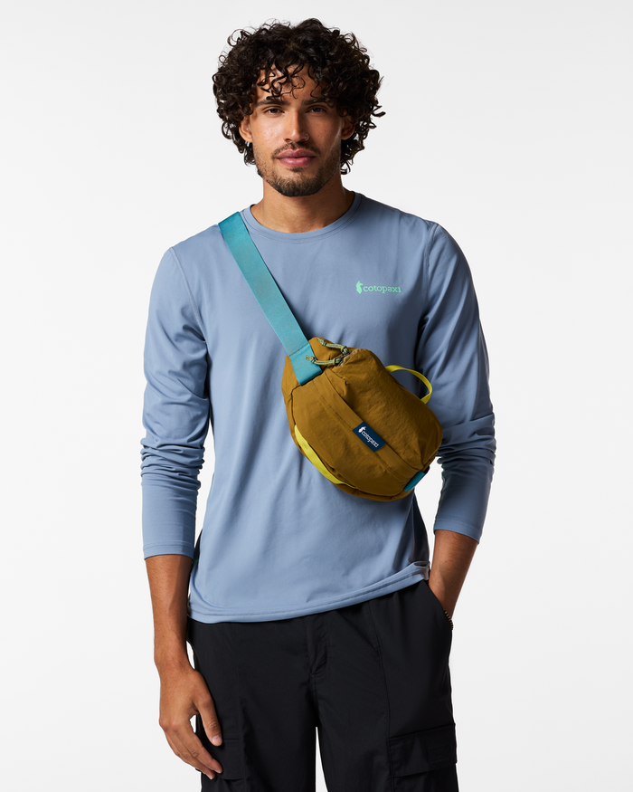 Man wearing a blue long-sleeve shirt and black pants, sporting the brown Empacable 3L Sling by Cotopaxi with a turquoise strap.