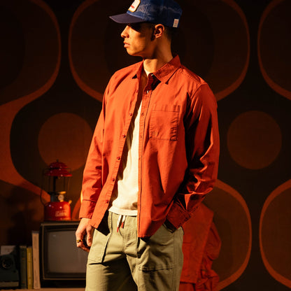 Topo Designs Men's Desert Shirt in orange, worn by a man with a blue cap and olive pants in a retro-themed room.