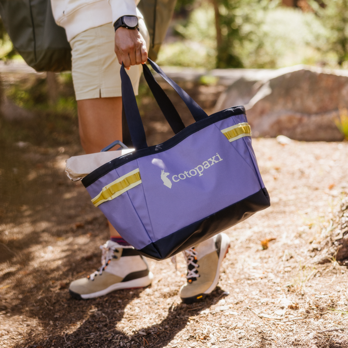 Person carrying a purple Cotopaxi Allpa 30L Gear Hauler Tote with yellow accents while walking outdoors, wearing hiking boots.