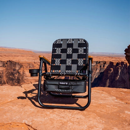 PARKIT Voyager Outdoor Chair on a canyon edge. Black & gray patterned straps, cupholder, wooden armrests, with scenic desert background.