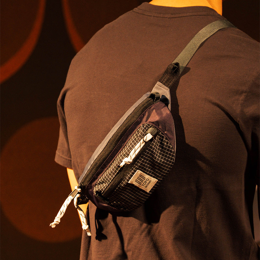 Mountain Waist Pack | Topo Designs on a person's back, dark color with grid pattern and logo, worn over the shoulder.