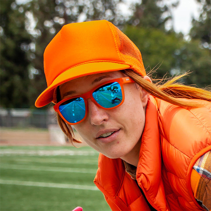 Woman wearing orange goodr Donkey Goggles sunglasses with blue mirrored lenses, an orange trucker hat, and vest. Track in background.