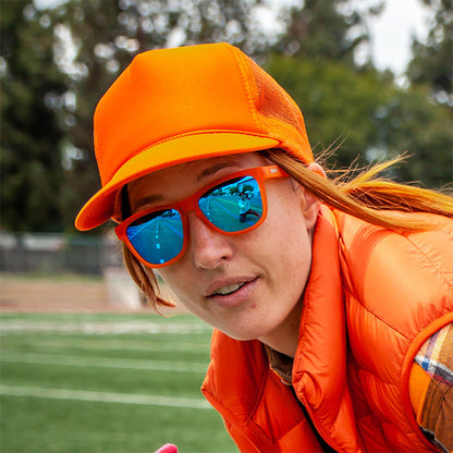 Woman wearing orange goodr Donkey Goggles sunglasses with blue mirrored lenses, an orange trucker hat, and vest. Track in background.