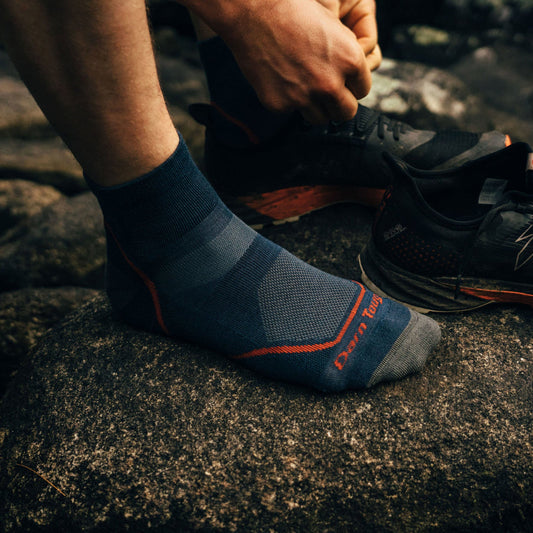 A person wearing blue and gray Darn Tough Men's Light Hiker Quarter Socks with orange accents, lacing hiking shoes on a rock.