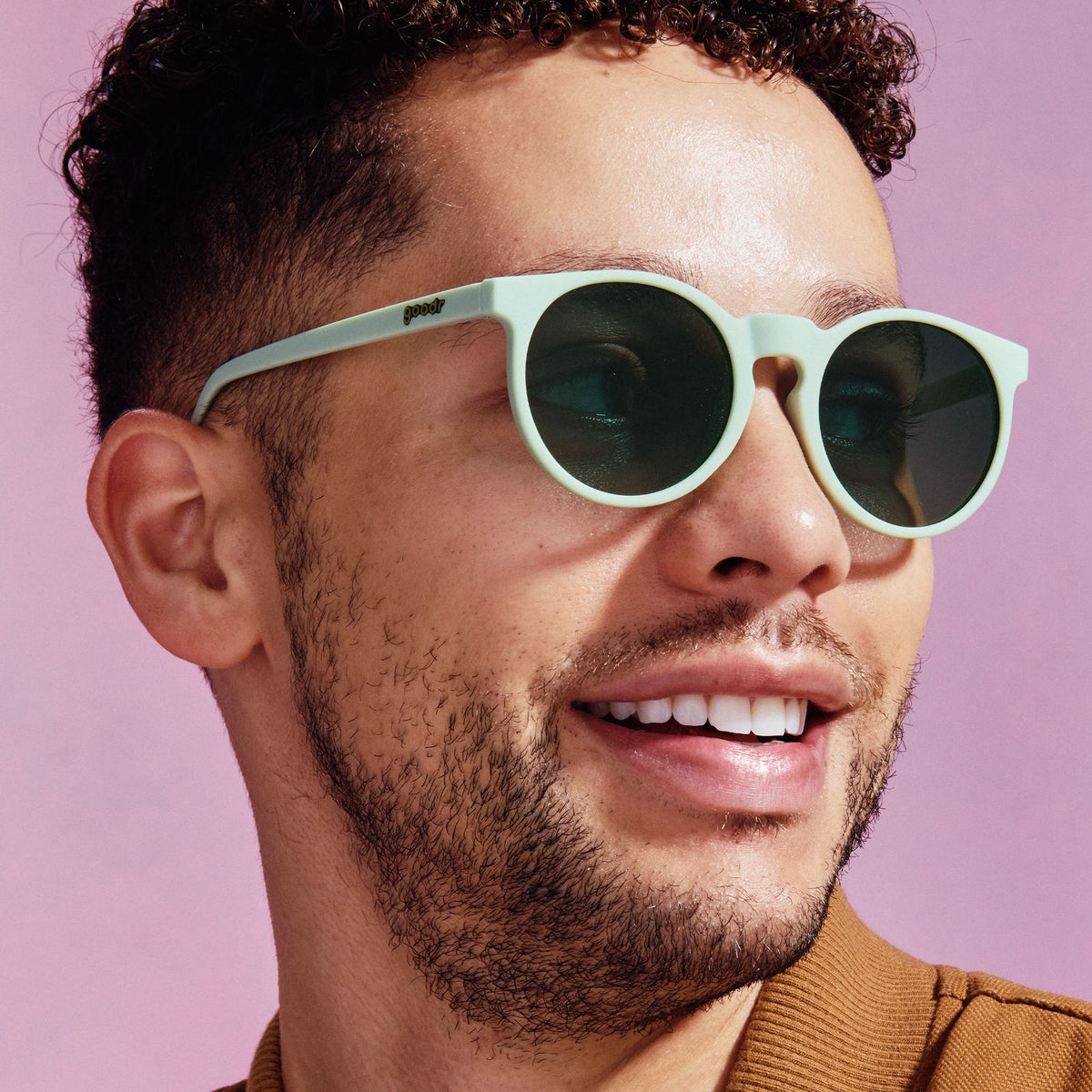 Man with curly hair and beard smiling, wearing Incognito Plant Parent goodr light mint green round sunglasses with dark lenses on a pink background.