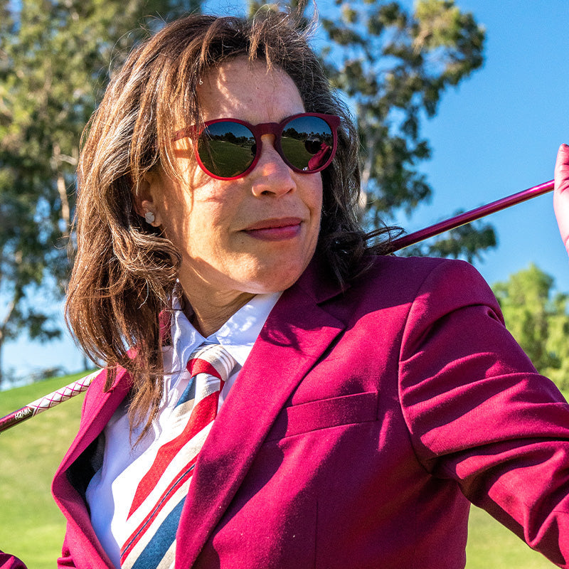 Smiling woman in burgundy blazer and tie wearing red goodr "I'm Wearing Burgundy?" round sunglasses while holding a golf club.