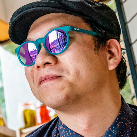Man in black cap wearing 'I Pickled These Myself | goodr' teal sunglasses with purple mirrored lenses, looking left.