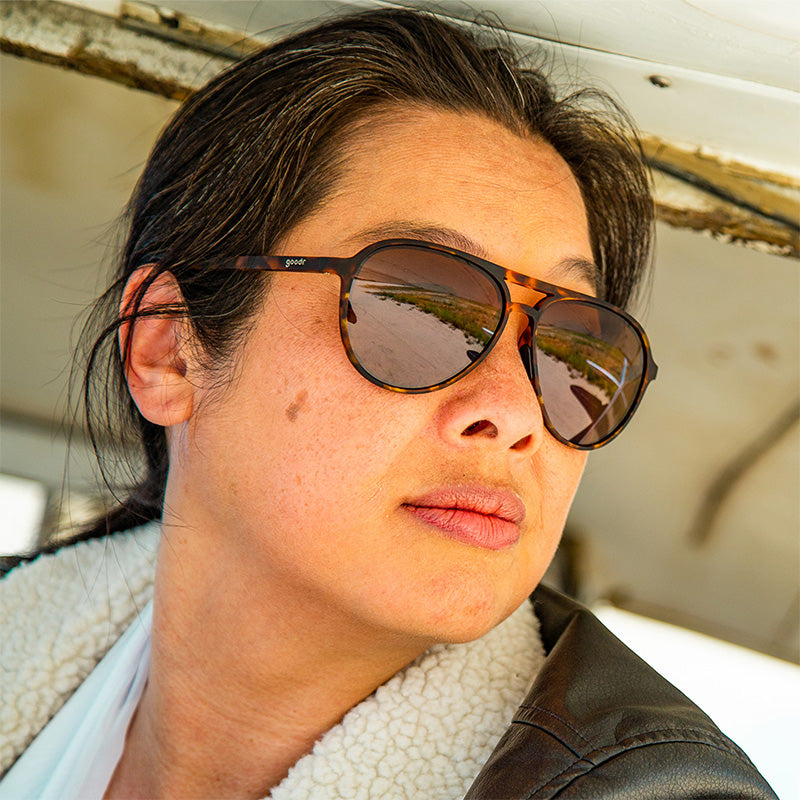 A person in a shearling jacket wearing goodr 'Amelia Earhart Ghosted Me' tortoise-shell sunglasses reflecting an outdoor path.