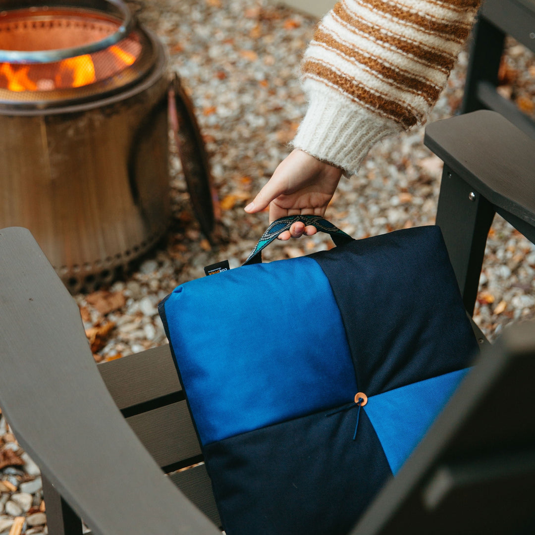 Hand placing a blue and black Wanderer 'QUAD' Outdoor Cushion from Media Mercantile on a chair by a fire pit.