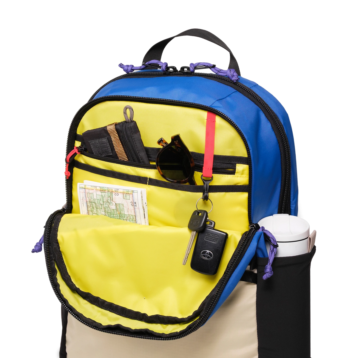 Topo Designs Commuter Backpack in blue and tan with an open front pocket showing internal organization, sunglasses, keys, map, and a water bottle in the side pocket.