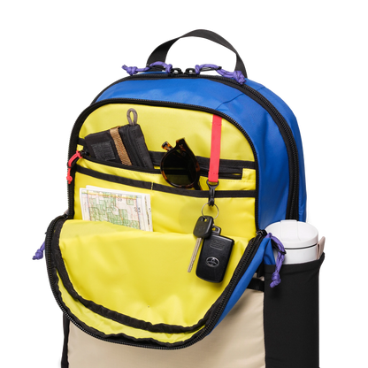 Topo Designs Commuter Backpack in blue and tan with an open front pocket showing internal organization, sunglasses, keys, map, and a water bottle in the side pocket.