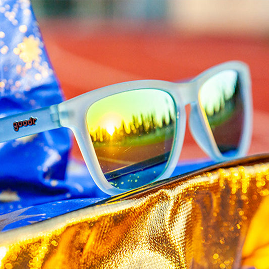 goodr 'Sunbathing with Wizards' sunglasses with mirrored lenses reflecting a sunset, resting on gold & blue fabric.