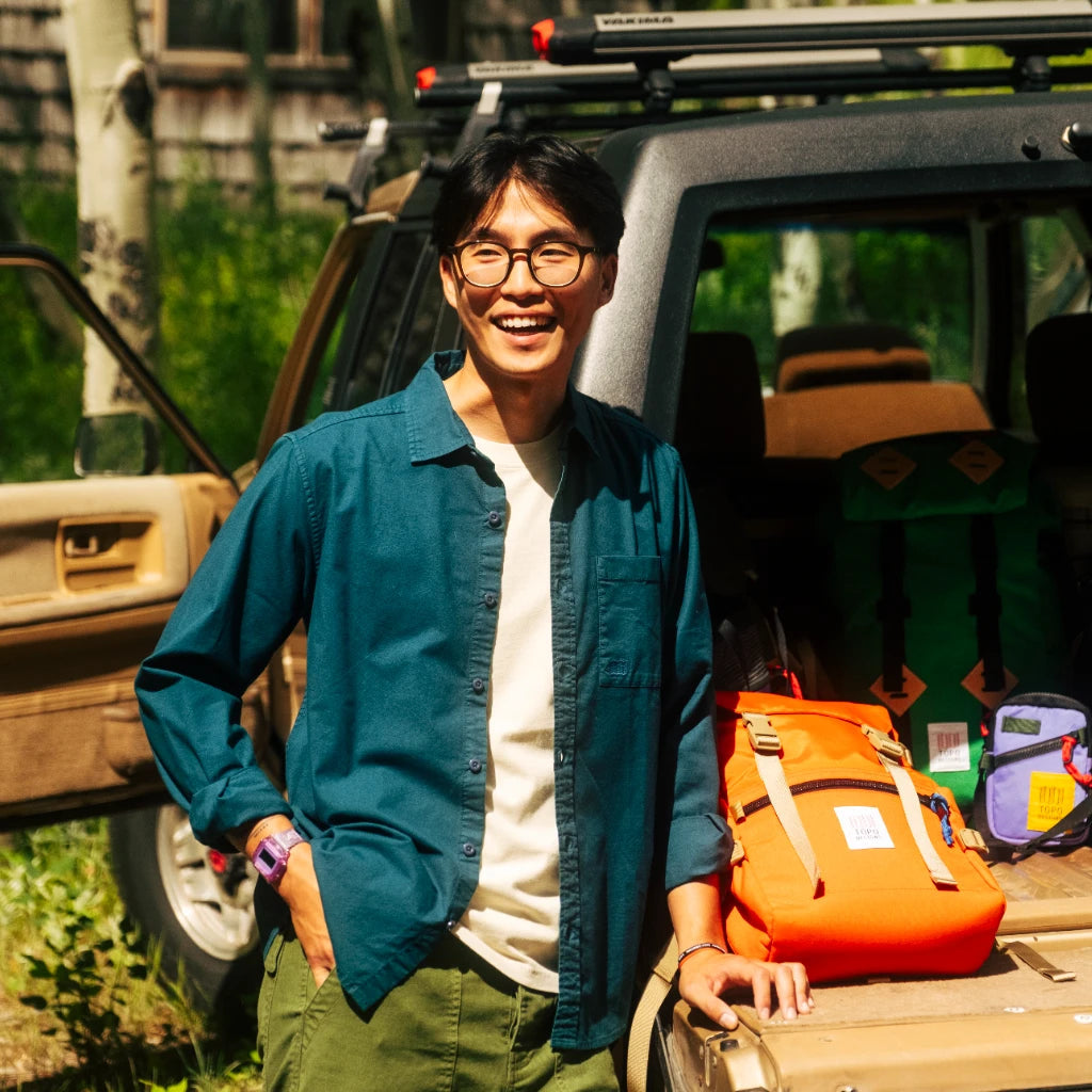 Topo Designs Men's Desert Shirt in teal, worn by a smiling man next to an SUV with colorful backpacks outdoors.