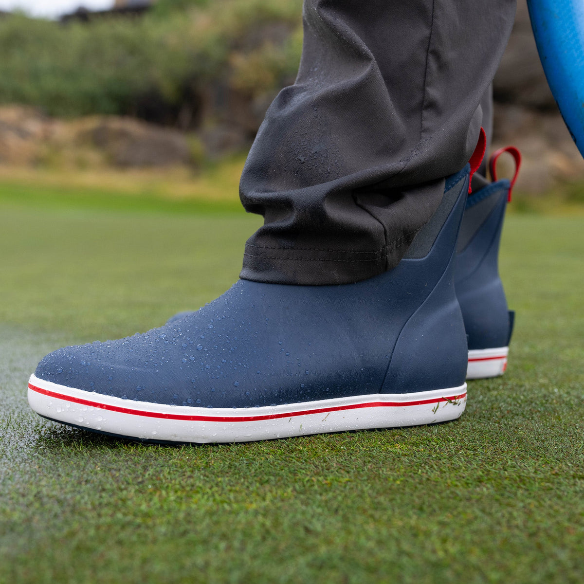XTRATUF Men's Ankle Deck Boot Sport in Stormy Blue, with white sole & red stripe, standing on wet green grass.