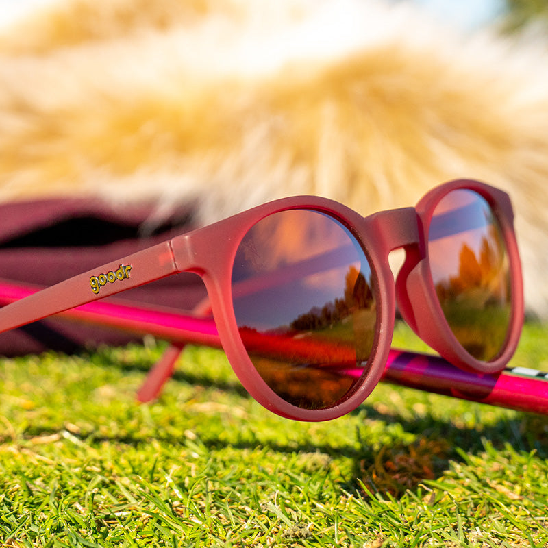 I'm Wearing Burgundy? goodr sunglasses on green grass, reflecting trees and sky in their lenses, with a blurred furry background.