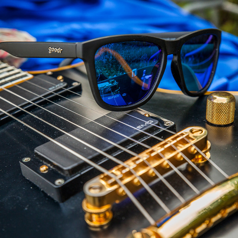 Mick and Keith's Midnight Ramble goodr sunglasses with blue reflective lenses on a black electric guitar.
