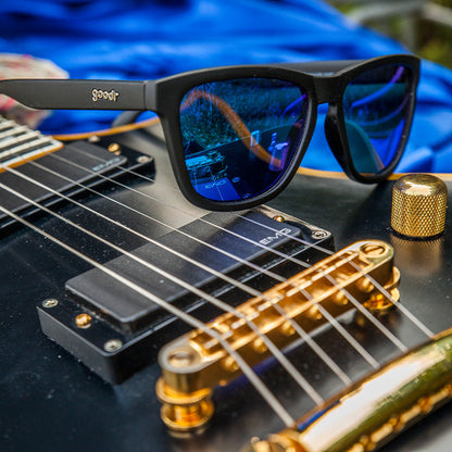 Mick and Keith's Midnight Ramble goodr sunglasses with blue reflective lenses on a black electric guitar.